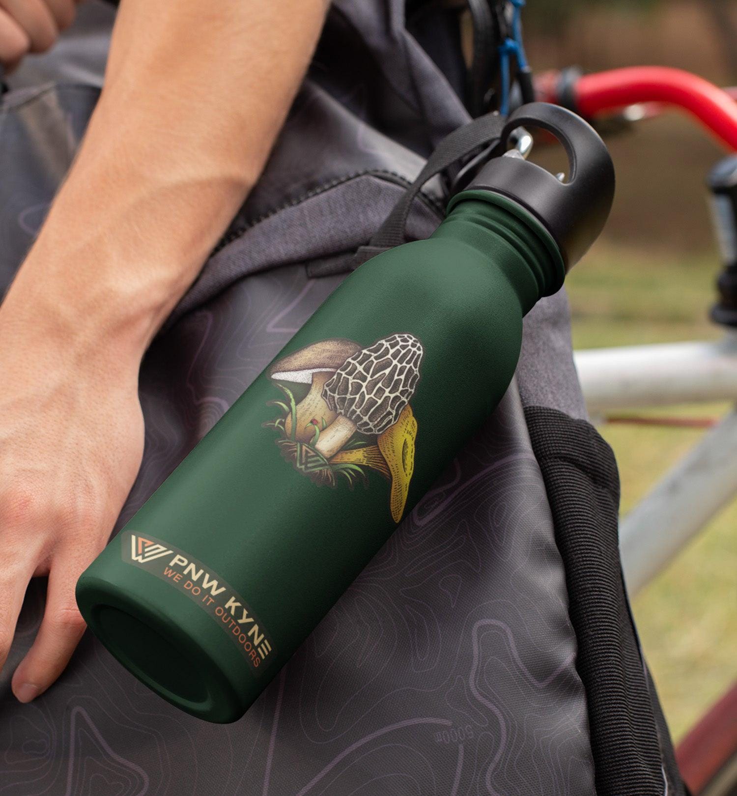 Green water bottle with a morel mushroom sticker on it, held by a person against a blurred Pacific Northwest outdoor background