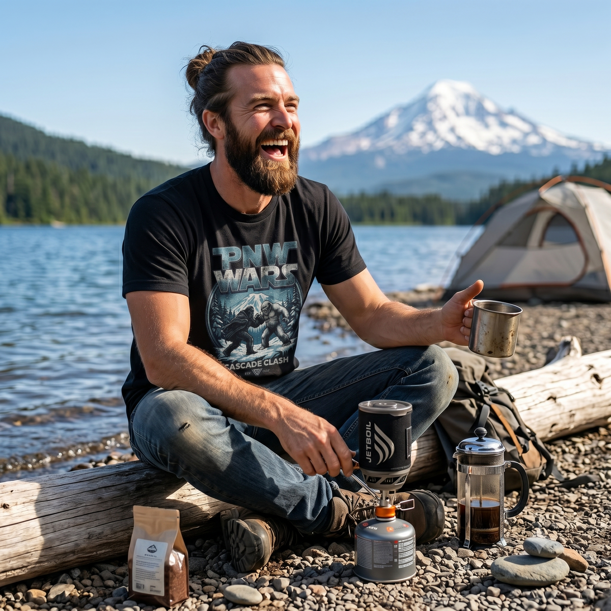 Man sitting by a lake wearing a PNW Wars Cascade Clash shirt, holding a coffee cup, and camping gear around him.